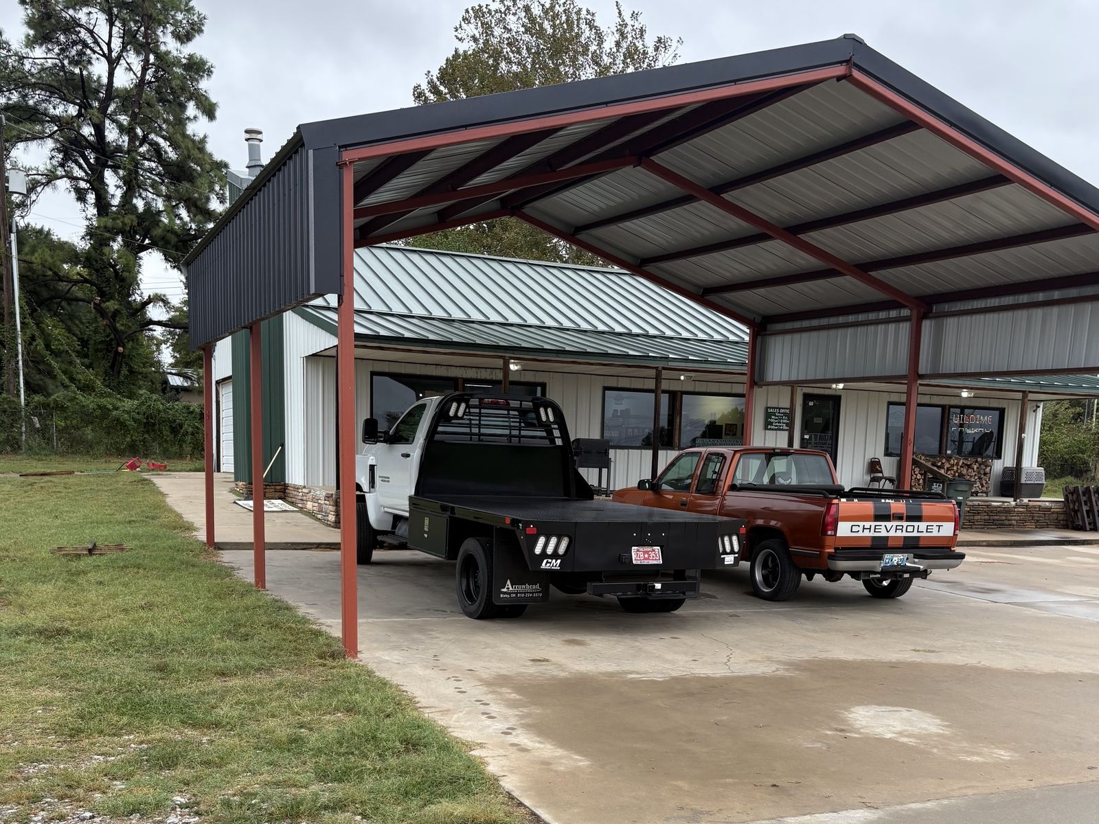 Open pole barn with red posts and a charcoal metal roof, a flatbed and pickup parked under it