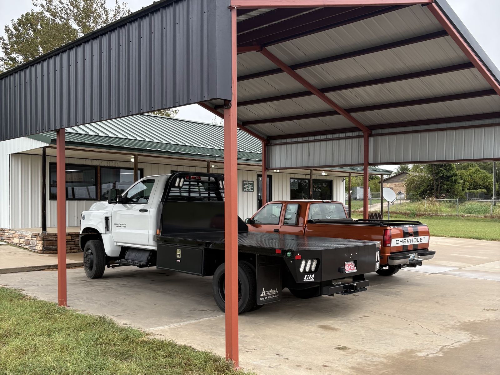 Pole barn with red posts and a charcoal upper wall, a flatbed truck and pickup parked under it